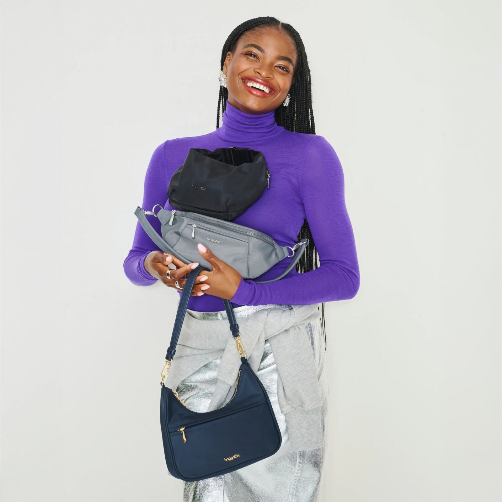 Smiling woman in a purple top holding a selection of black, gray and navy Baggallini bags.