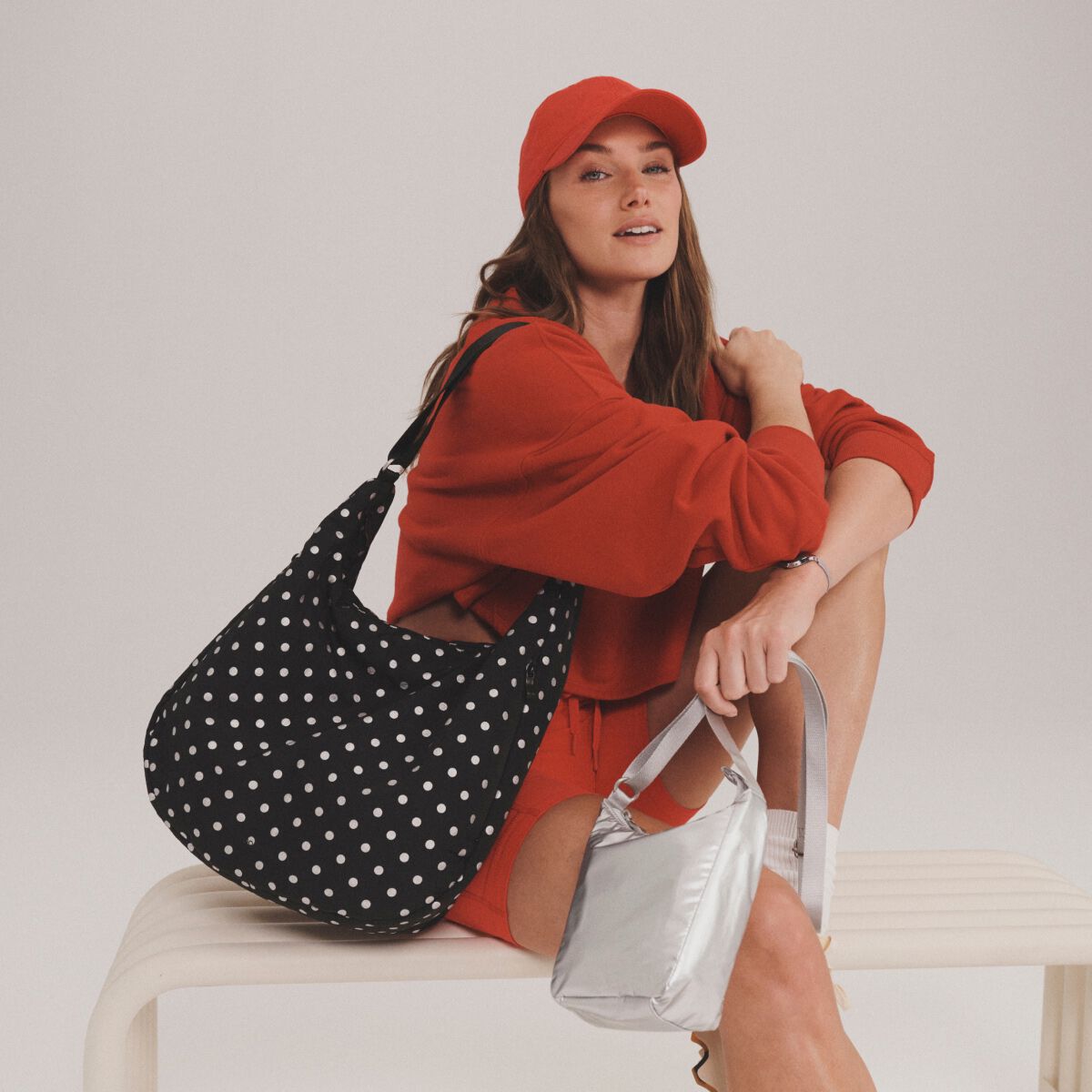 Model in red holding a black polka dot Baggallini bag with a silver crossbody bag beside her.